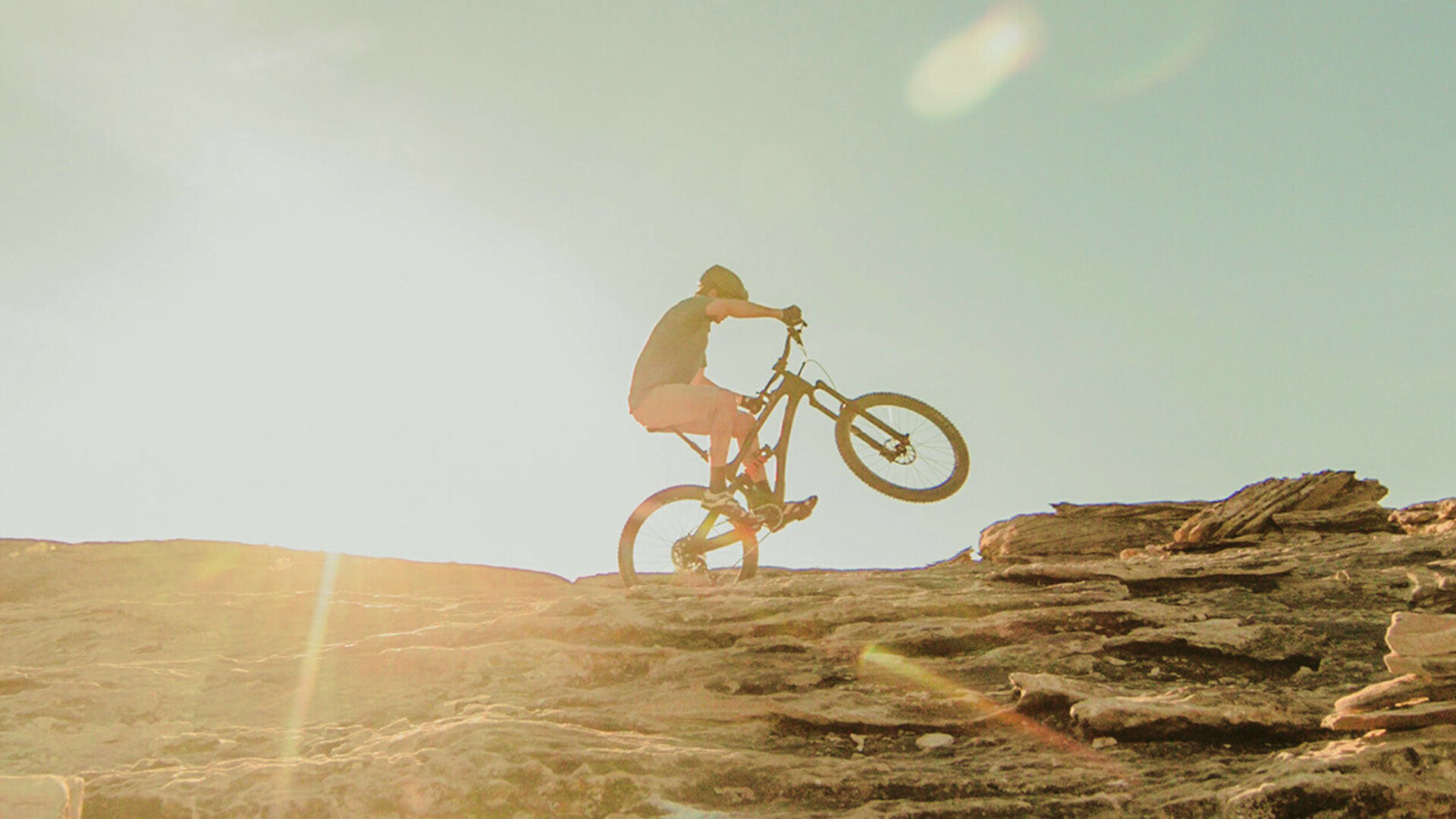 Mountainbiker fietst een rotsachtige helling op bij zonsondergang, met zichtbare zonnestralen op de achtergrond.