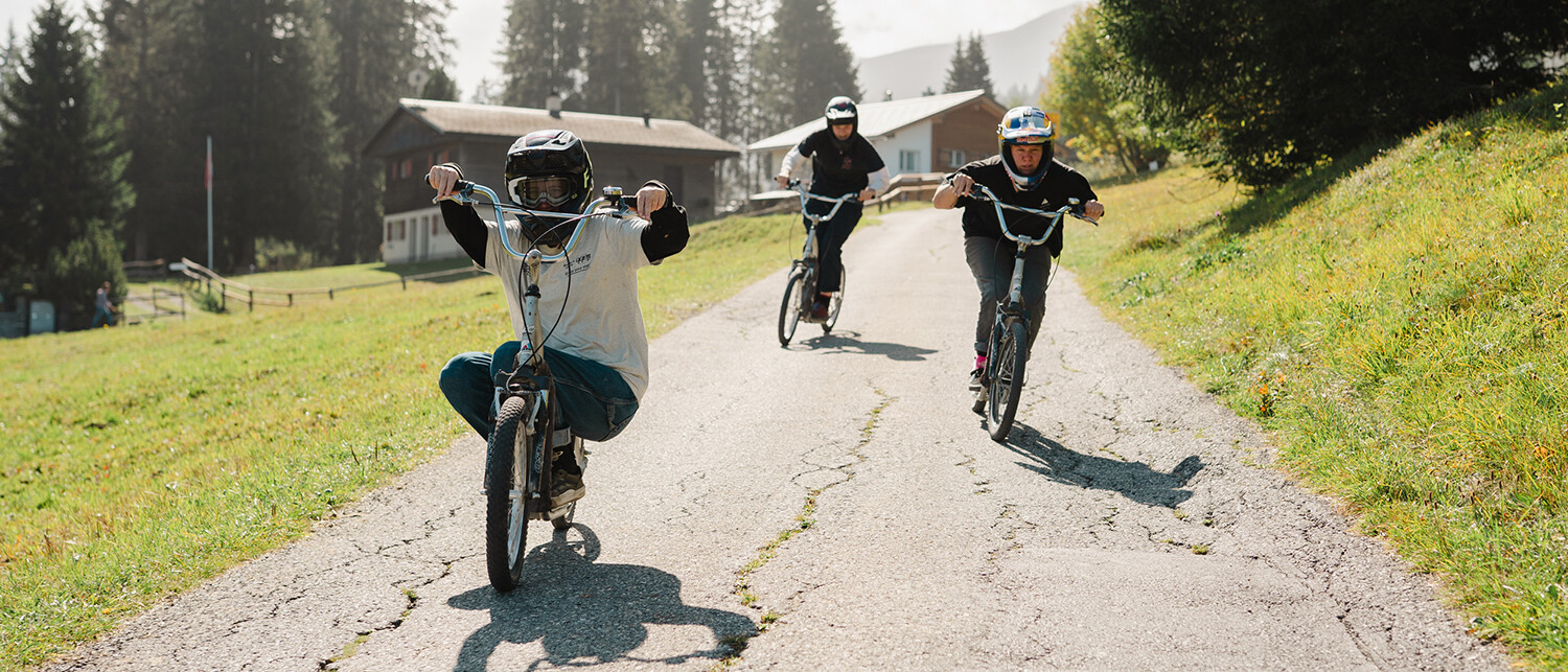 Three cyclists wearing helmets riding down a paved rural path, surrounded by grass and trees, with houses in the background.