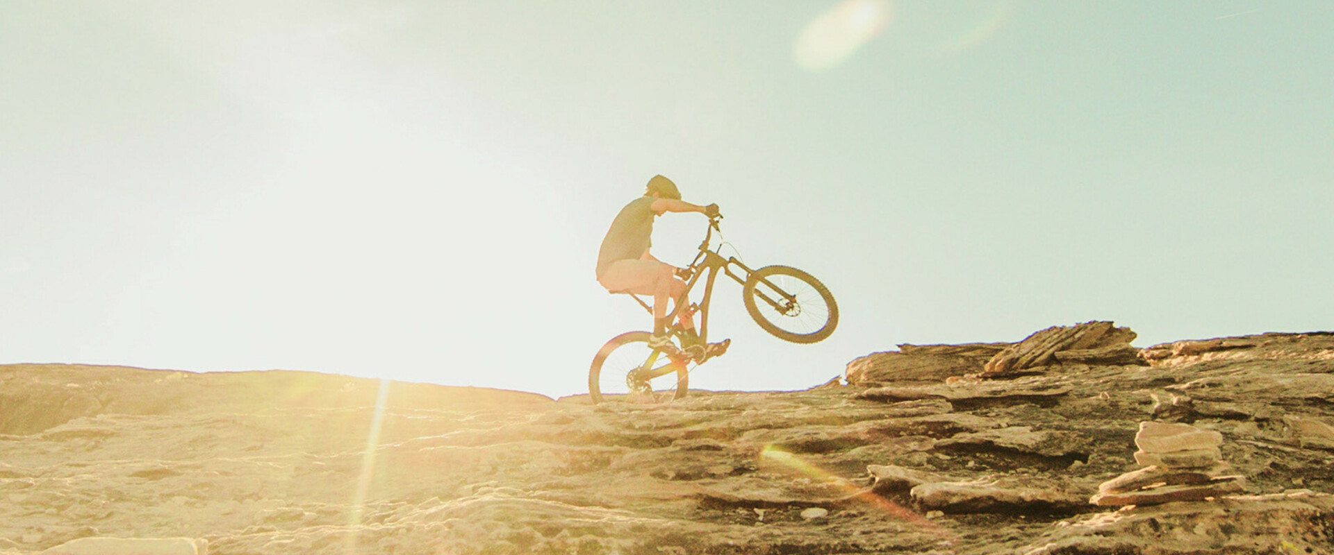 Mountainbiker fietst een rotsachtige helling op bij zonsondergang, met zichtbare zonnestralen op de achtergrond.