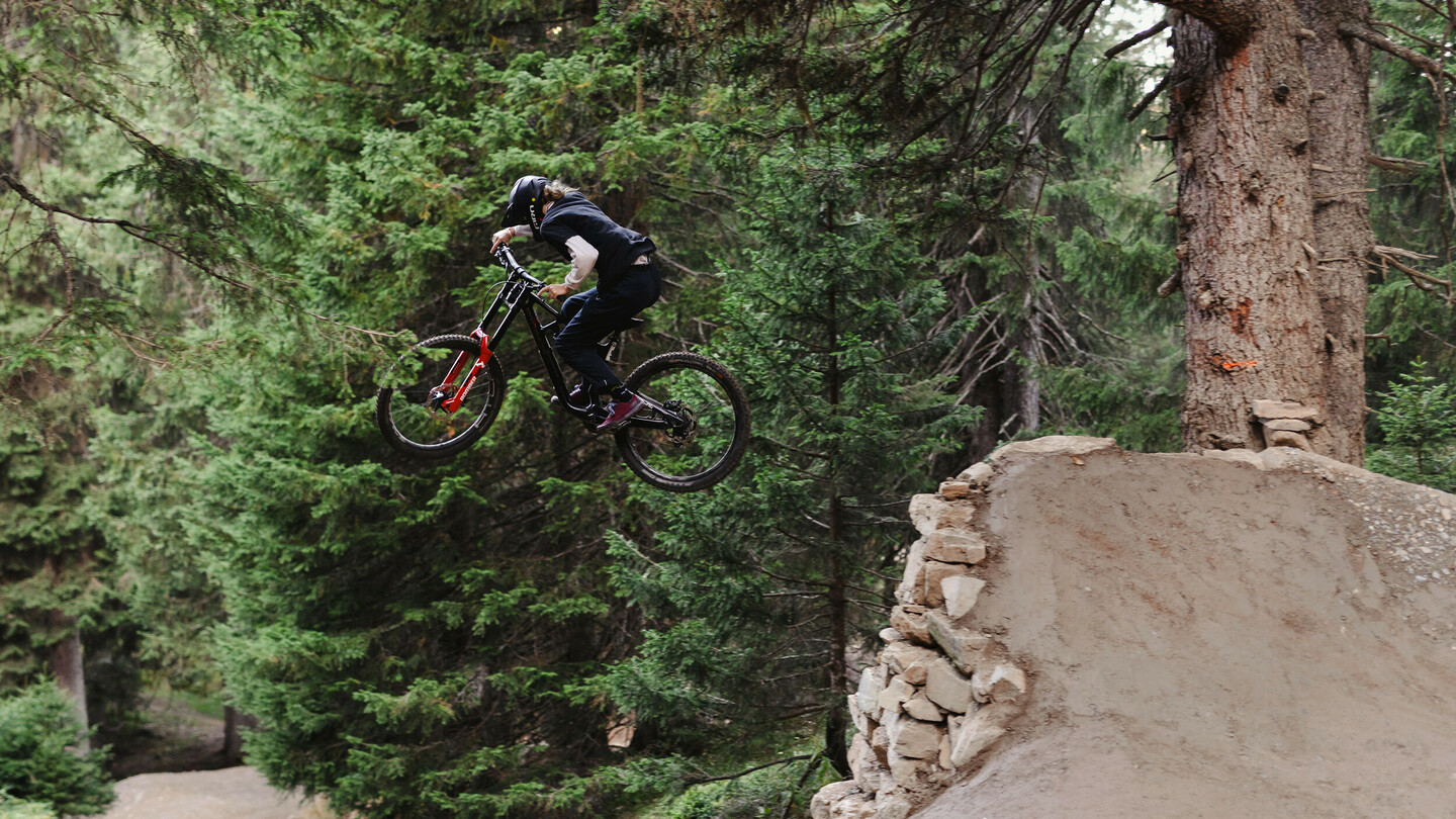 Een mountainbiker springt met zijn fiets van een ramp in een dicht bos. De rijder draagt een helm en donkere kleding terwijl hij over een grote aarden schans vliegt.