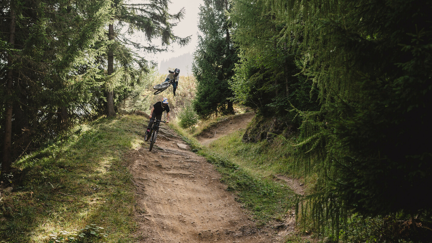 Een mountainbiker voert een hoge luchttruc uit terwijl een andere rijder een bosweg volgt. De scène is omgeven door hoge bomen en weelderige vegetatie.