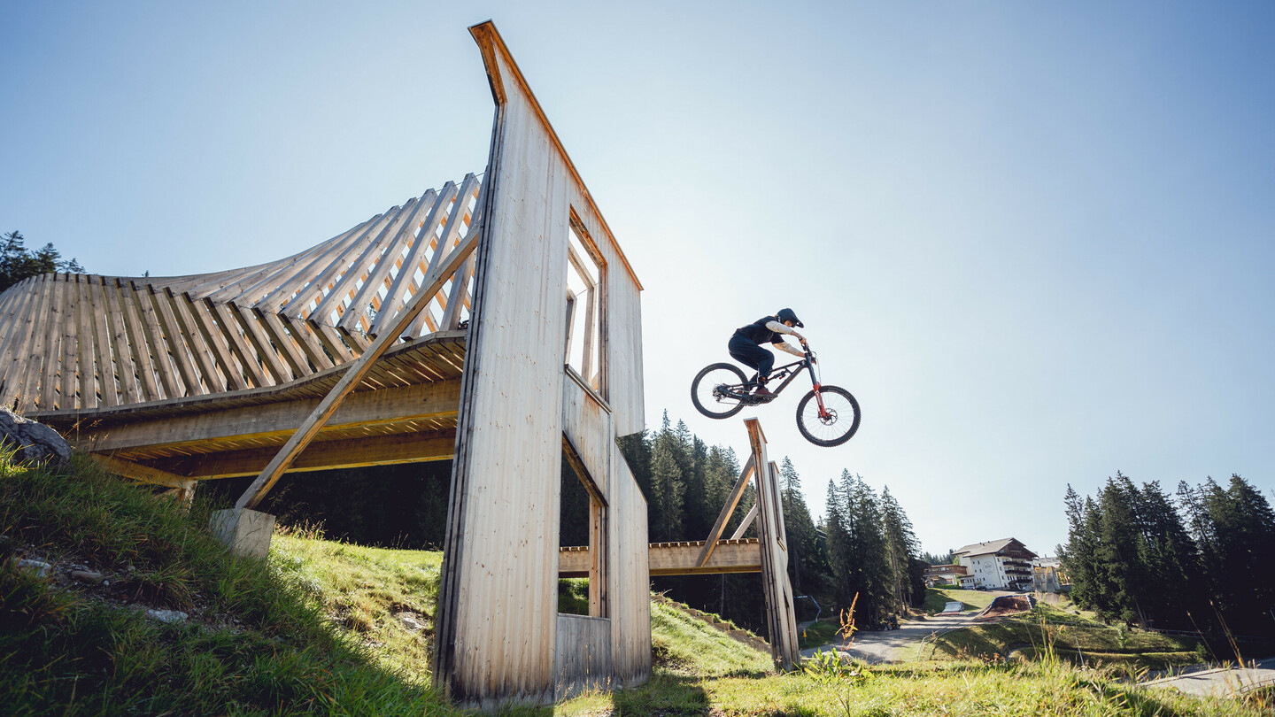 A mountain biker jumps over a wooden ramp in a bike park. The massive wooden structure stands out against the clear sky and surrounding nature.