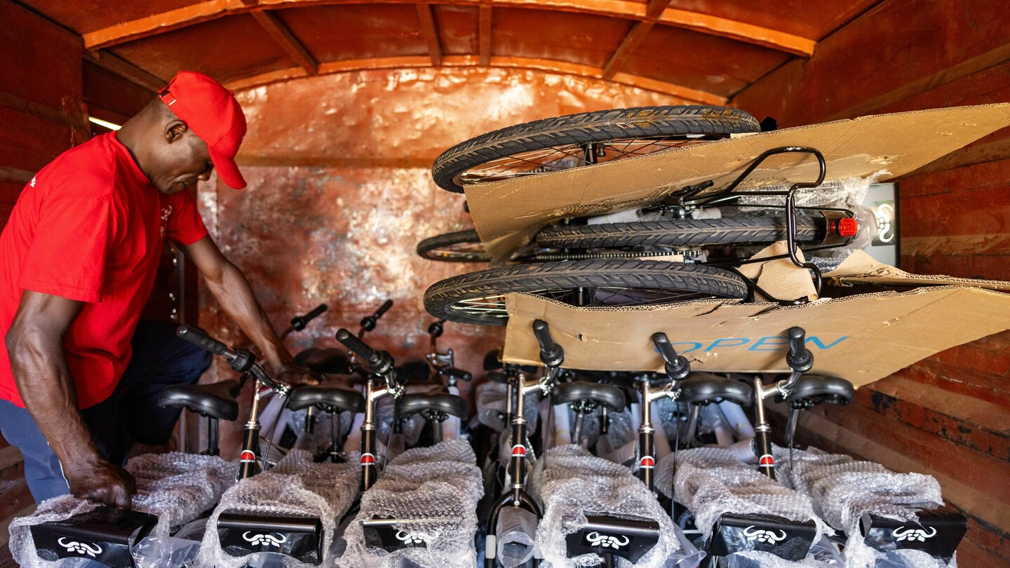 A man in a red shirt and cap loading bicycles into a delivery van. The bicycles are partially packaged and stacked. The interior of the van has a rusty tone.