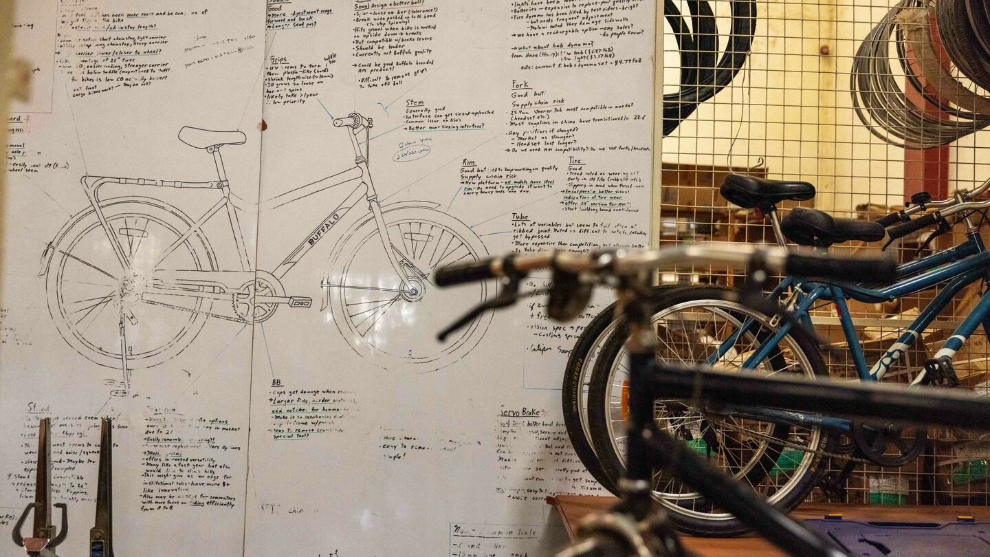 Tekening van een fiets op een muur, omringd door handgeschreven notities en technische details. Vage fietsen en onderdelen zijn zichtbaar op de voorgrond.