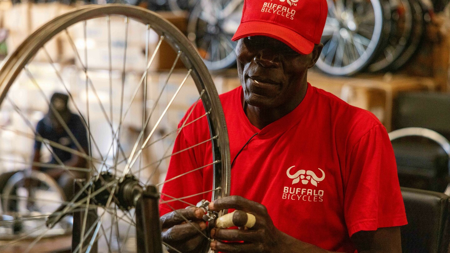 Een man in een rood shirt en pet met Buffalo Bicycles-logo werkt aan een fietswiel in een werkplaats. Op de achtergrond zijn gestapelde fietsen en dozen vaag zichtbaar.