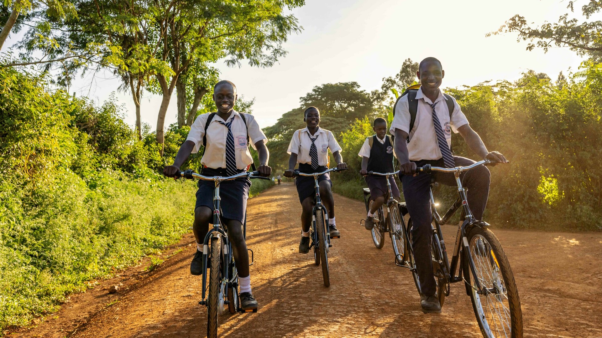 Groep scholieren in schooluniformen fietst over een onverharde weg.