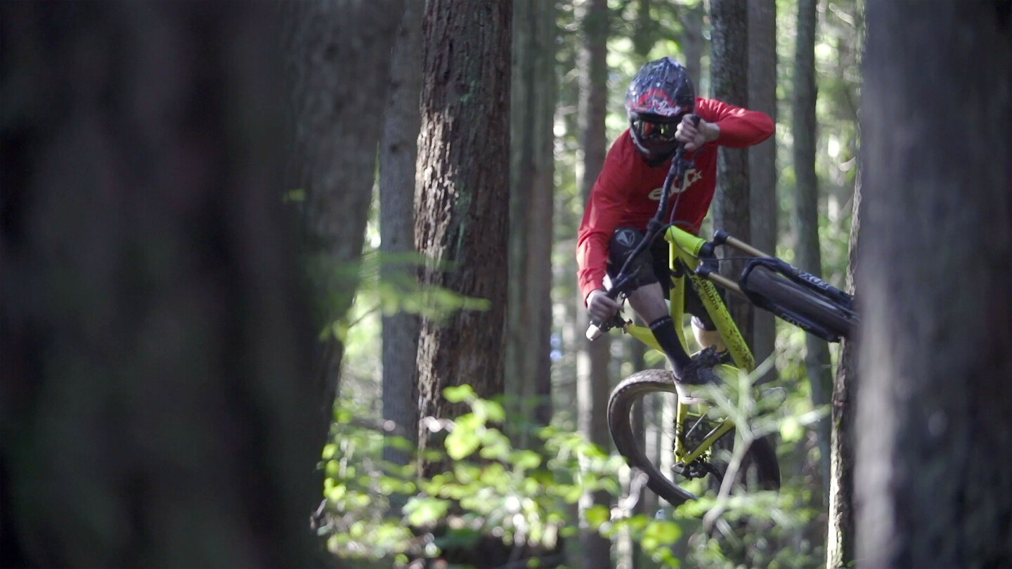 Mountainbiker met helm en rood shirt voert een sprong uit in een dicht bos, waarbij de fiets zijwaarts gekanteld is.