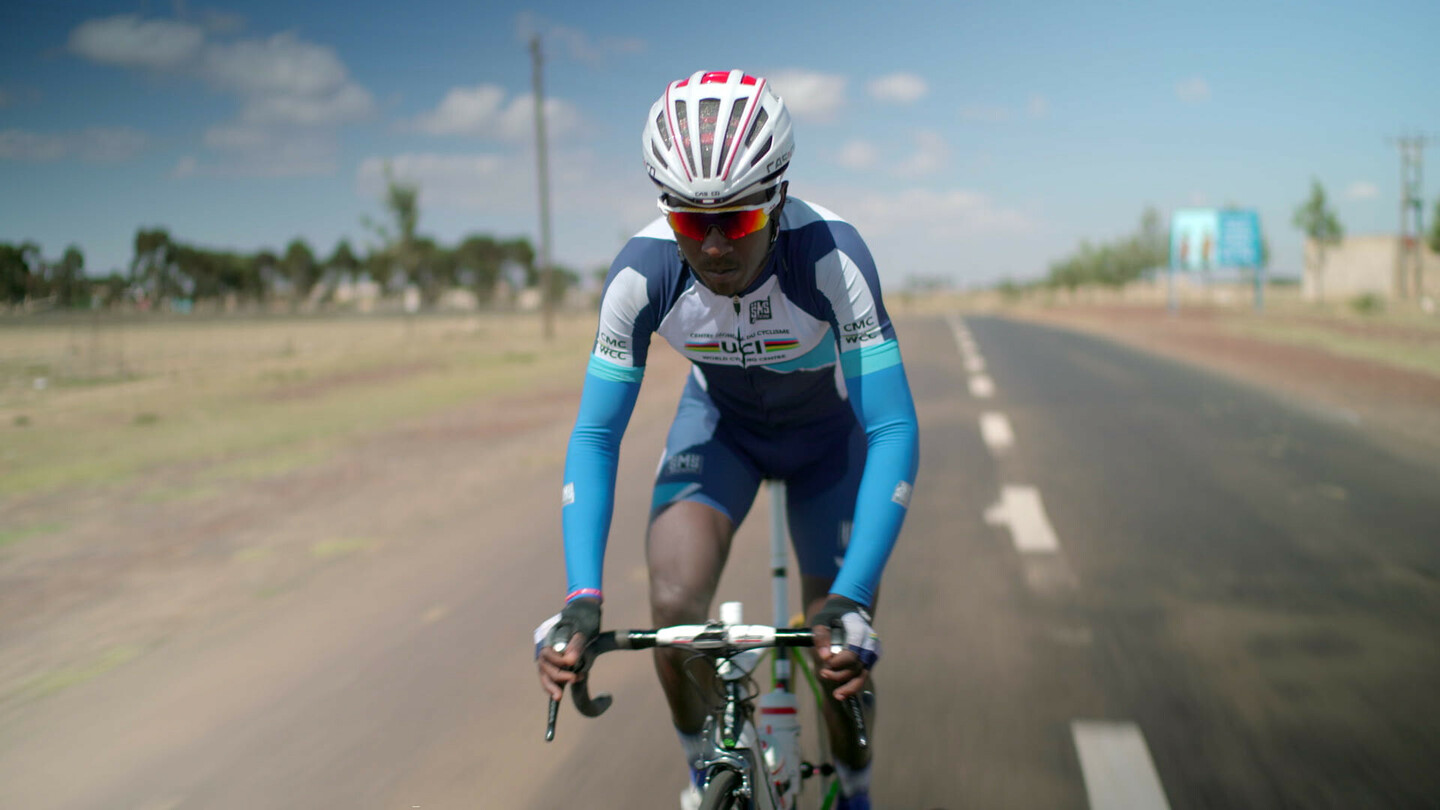 Biniam Girmay rijdt op een racefiets op een open weg, gekleed in een blauw-wit UCI-wielertenue en een helm.